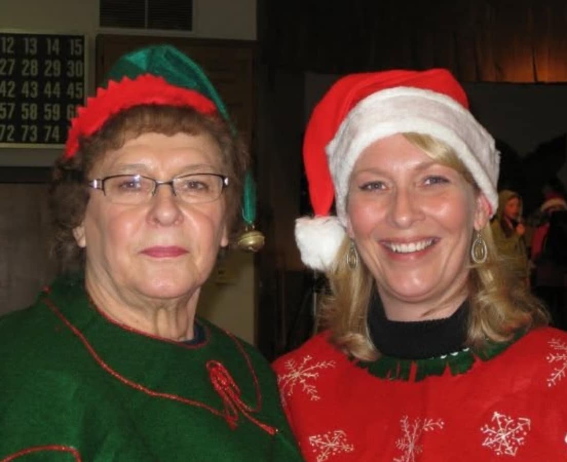 Mother in a green elf hat and daughter in a Santa hat, side by side at a Christmas party, both smiling
